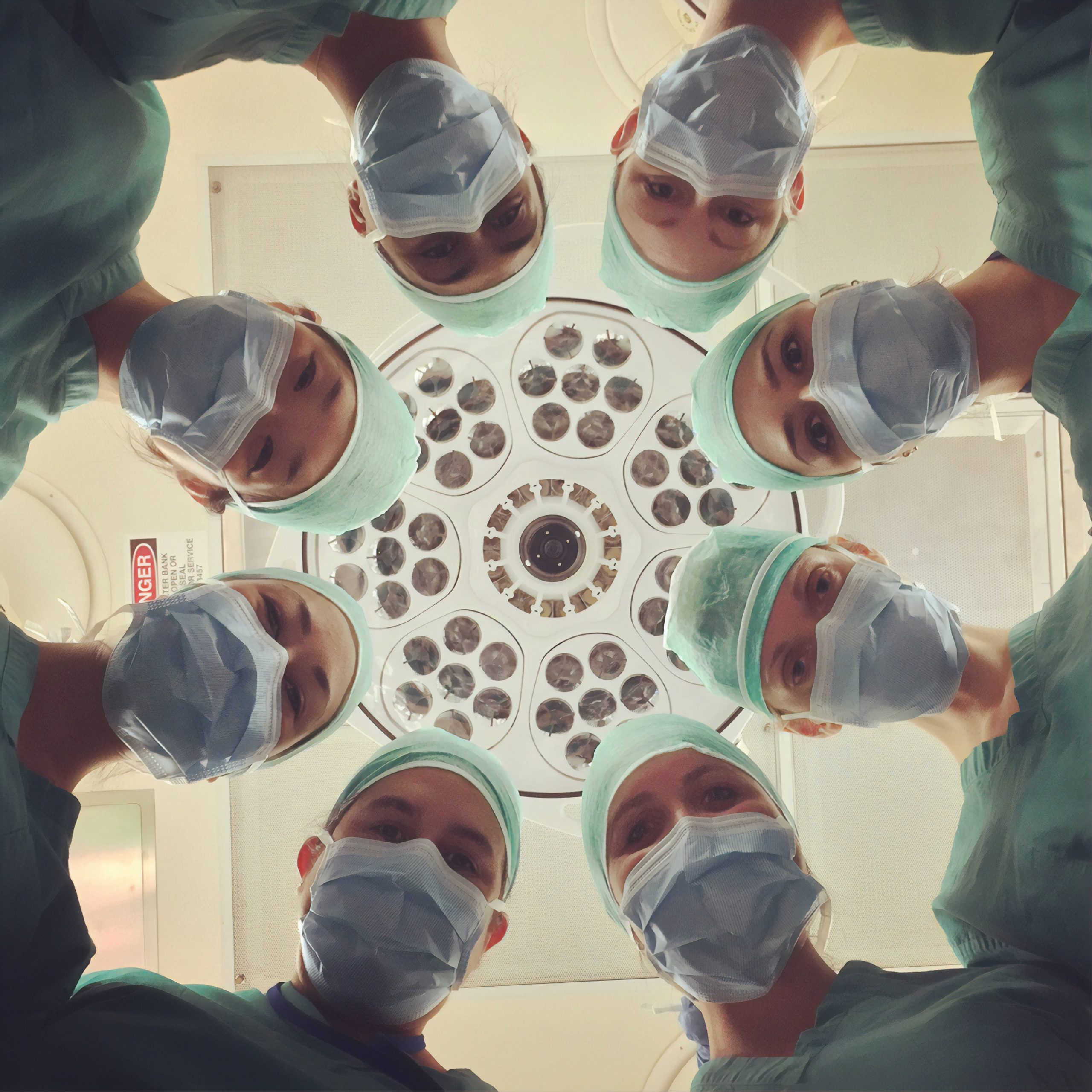 masked doctors in scrubs stand in a circle and peer down at the camera